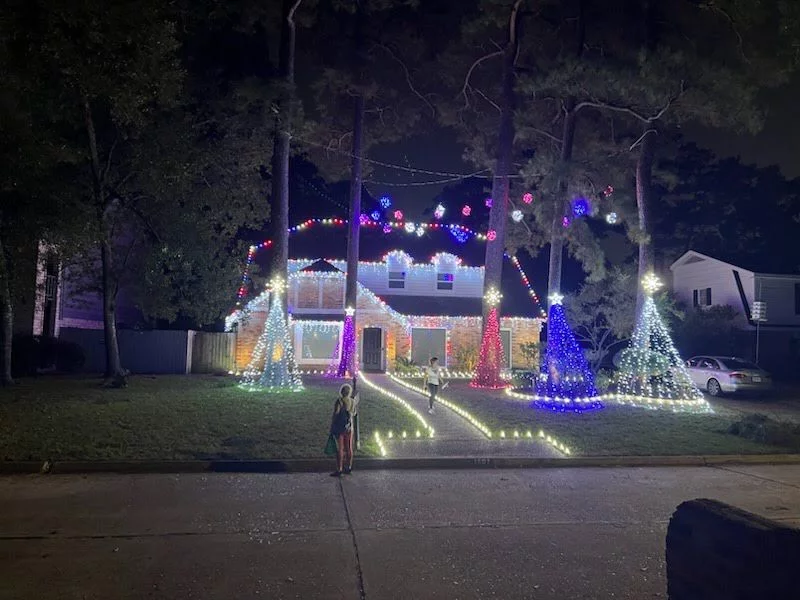 A house at night decorated with colorful lights, illuminated trees, and a glowing walkway, with a child standing in the foreground.