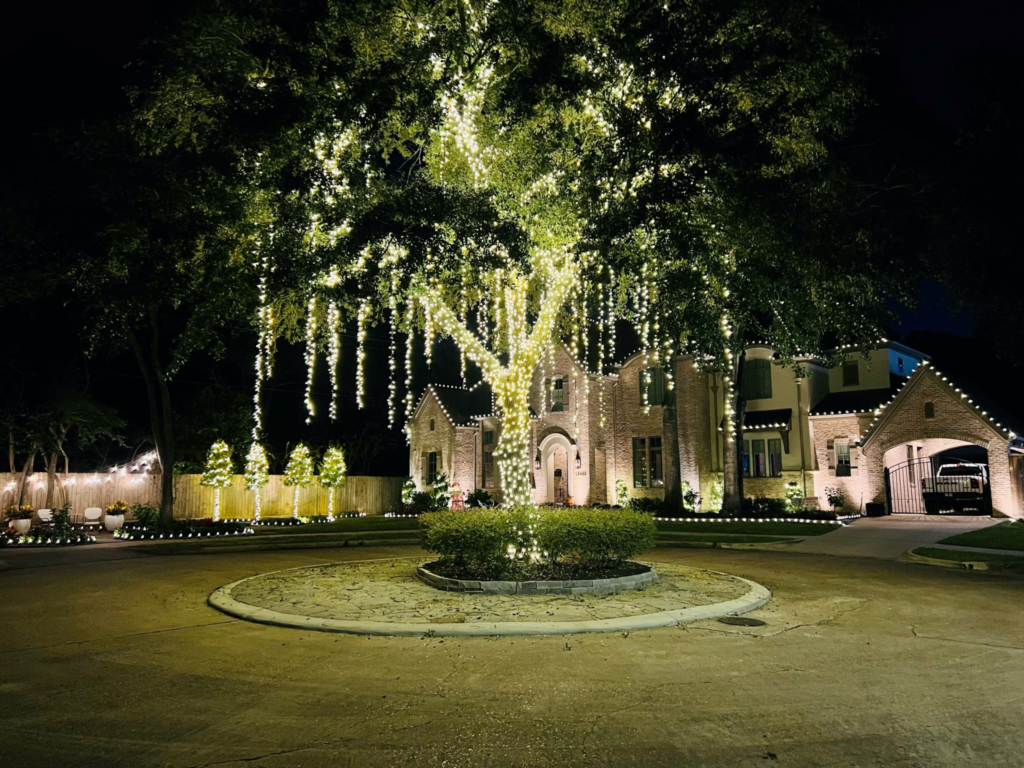 A large tree draped in cascading white lights stands in front of an illuminated brick house at night.
