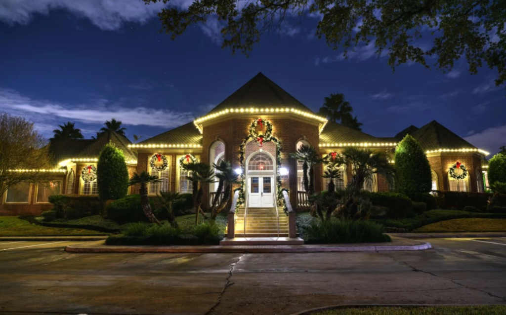 A large brick building at night is warmly lit with Christmas lights and wreaths, featuring a central entrance with steps and festive decorations.