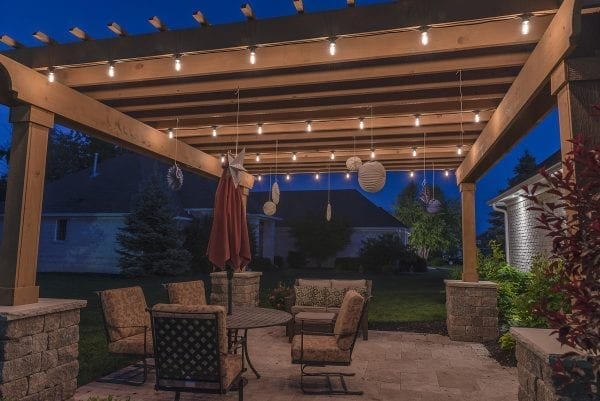A cozy backyard patio under a wooden pergola, softly lit with string lights at dusk and furnished with outdoor seating.