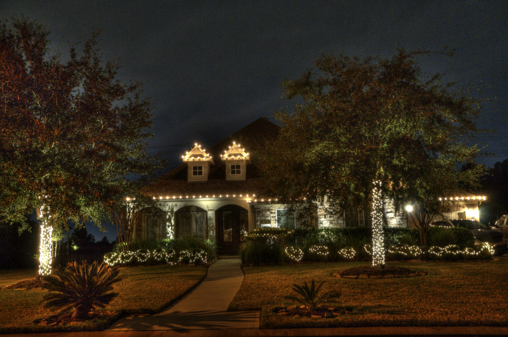 A charming house at night decorated with warm white lights on the roof, trees, and garden, creating a festive glow.