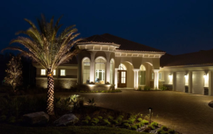 A large, elegant Mediterranean-style house illuminated at night with warm exterior lighting, arched windows, and a palm tree in the front yard.