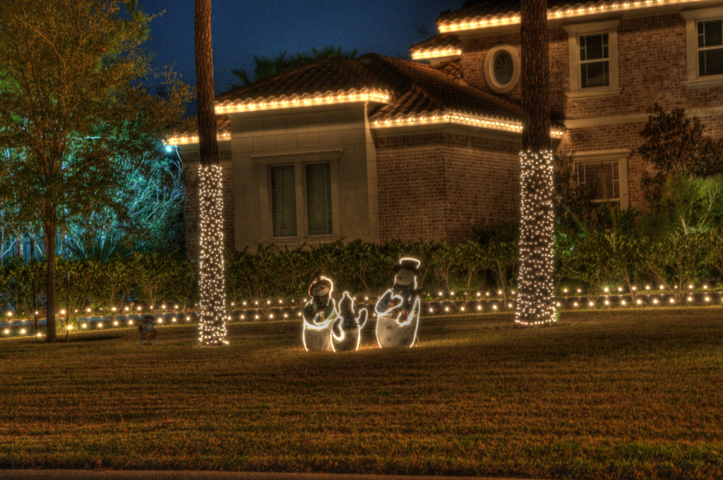 A brick house at night decorated with festive holiday lights, glowing palm trees, and illuminated snowman figures on the lawn.