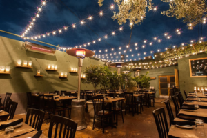 A cozy outdoor restaurant patio at night, warmly lit with string lights, candles, and heaters over neatly set tables.