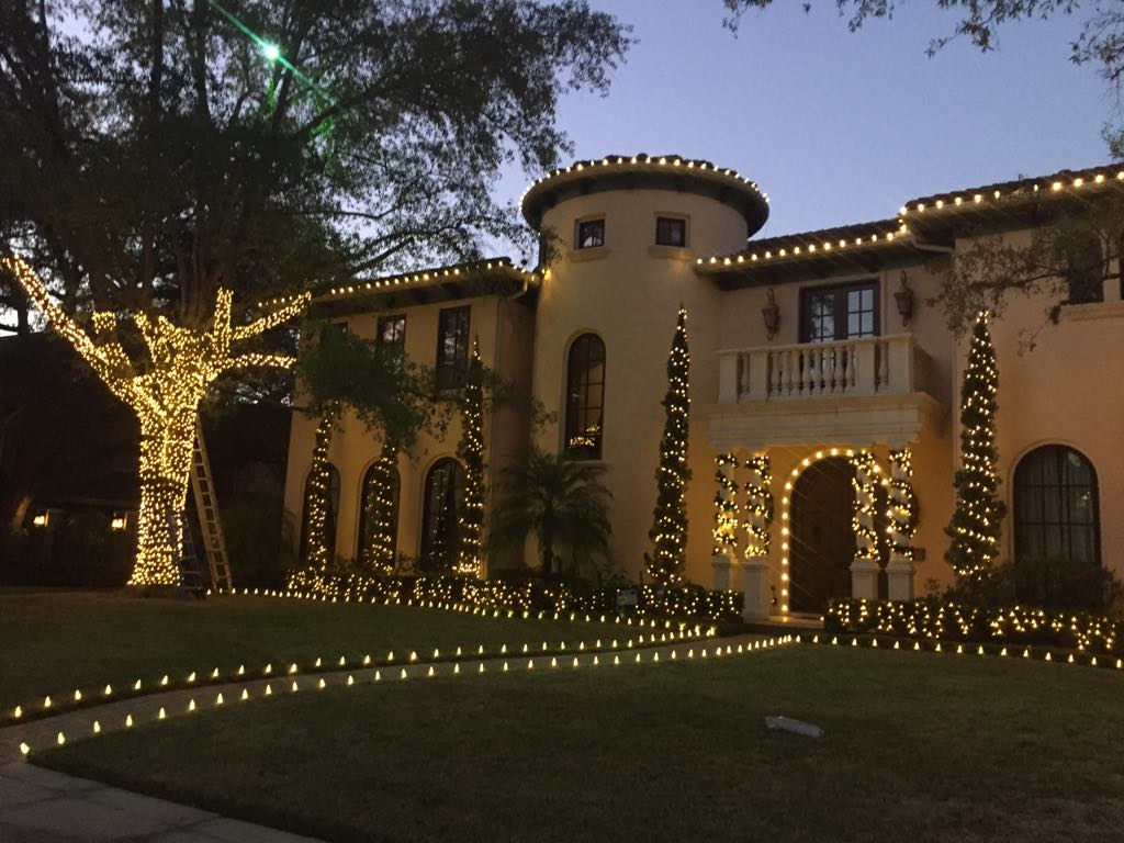 A Mediterranean-style villa at dusk, decorated with warm white holiday lights on the house, trees, and lawn.